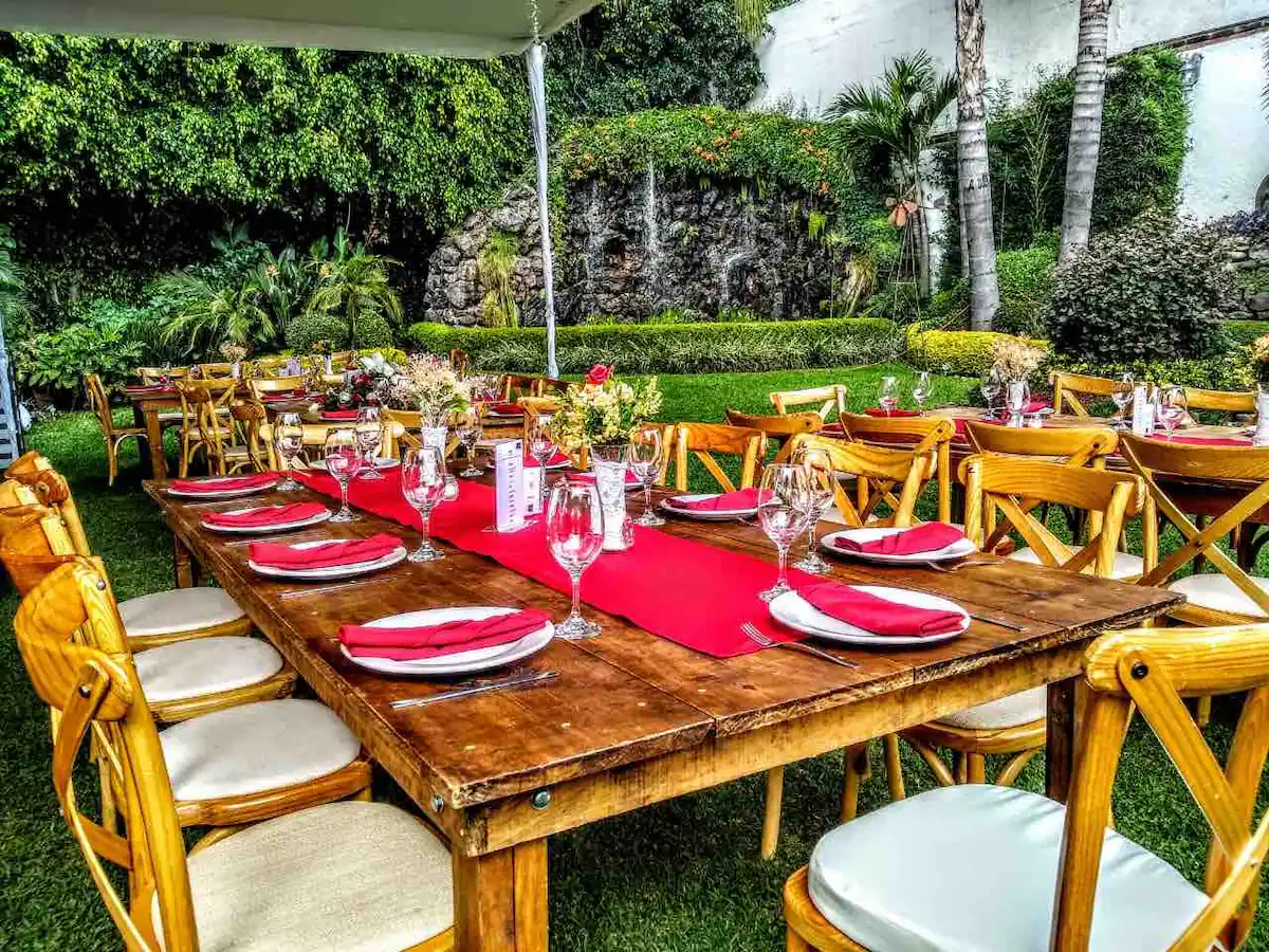Outdoor dining area with wooden tables and chairs, red table runners, and glassware, set in a lush garden with greenery and a waterfall feature.