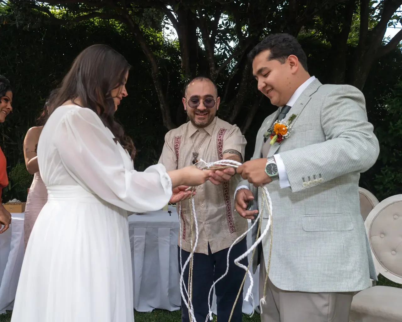 A wedding ceremony outdoors with a bride, groom, and officiant exchanging vows under a large tree.