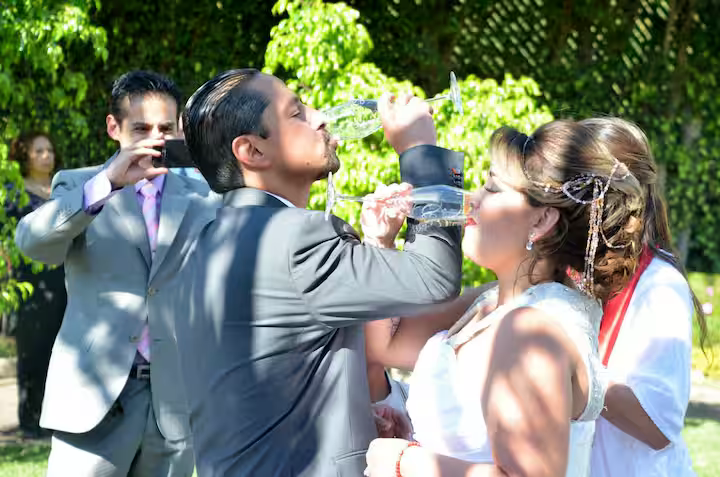 A bride and groom sharing a toast with champagne at their wedding reception outdoors, with guests in the background.