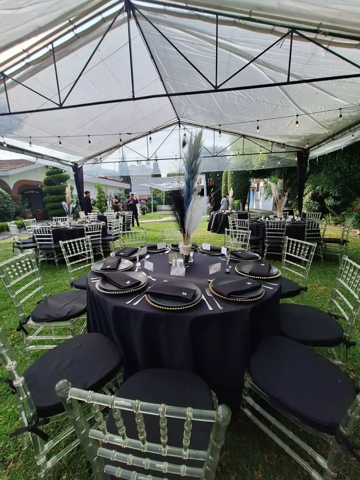 Elegant outdoor event setup with round tables covered in black tablecloths, black napkins with silver accents, and clear chairs with black cushions. Centerpieces feature tall vases with black and white feathers, under a white canopy decorated with st