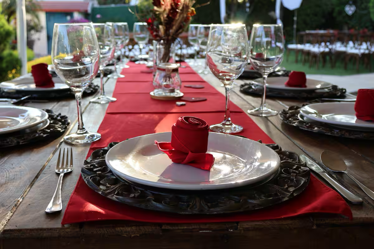 A formal dining table set outdoors with plates, wine glasses, silverware, and red napkins, featuring a red napkin rolled into a rose shape as a centerpiece.