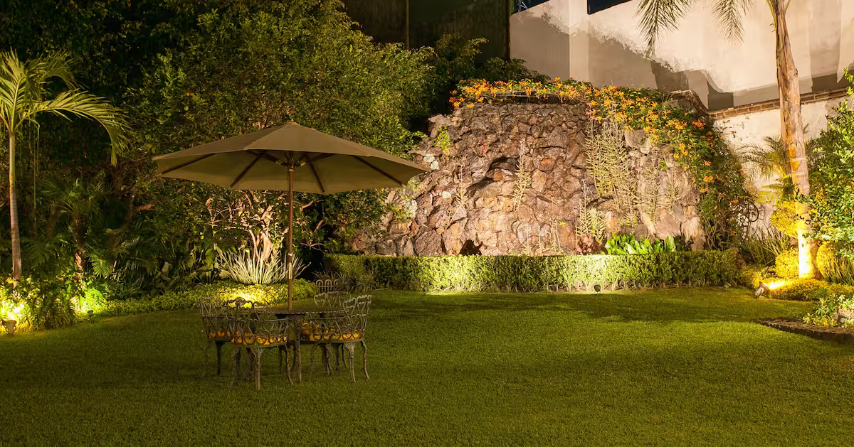 Night view of a landscaped backyard with a stone fountain, surrounded by lush greenery, illuminated by outdoor lighting, with a patio table and chairs with an umbrella.