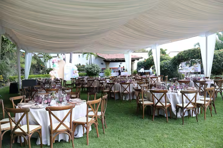 Outdoor wedding reception setup under a large white tent with multiple round tables, decorated with white tablecloths and floral centerpieces, surrounded by wooden chairs with beige cushions, on a grassy lawn.