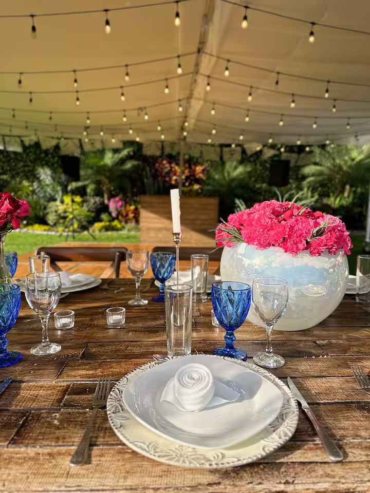 Set dining table with white plate, wrapped napkin, and silverware, decorated with pink flowers in a white vase, under string lights in an outdoor tent.