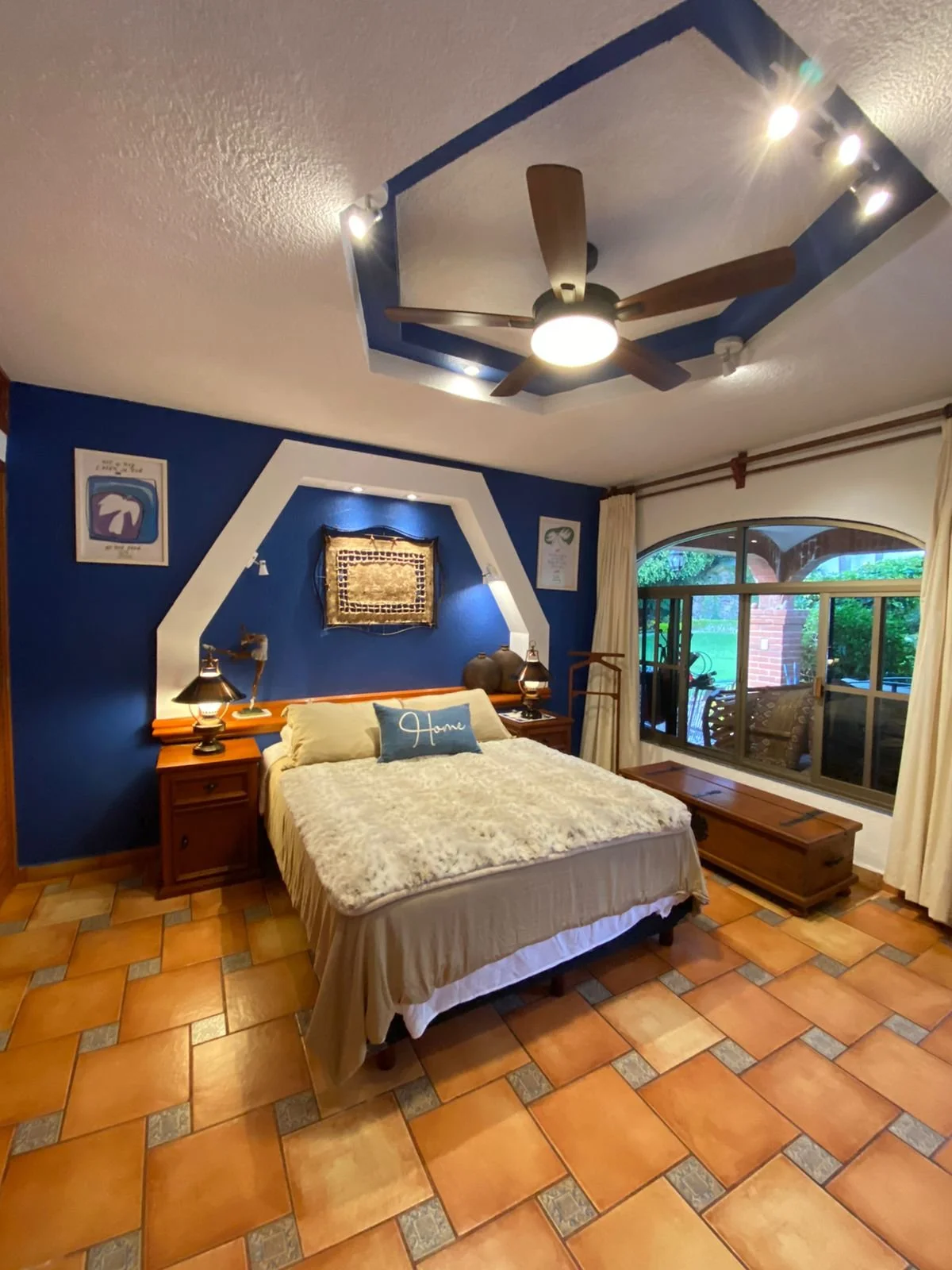 A cozy bedroom featuring a bed with a "Home" pillow, nightstands with lamps, a large window with curtains, a ceiling fan, and decorative wall art.