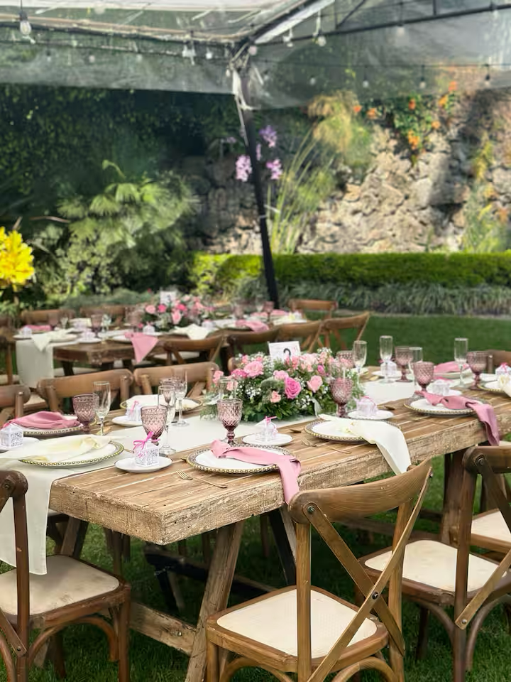 Garden outdoor party setup with rustic wooden tables, pink and white flower centerpieces, pink and white napkins, pink-tinted glasses, and wooden chairs amidst greenery and rocks