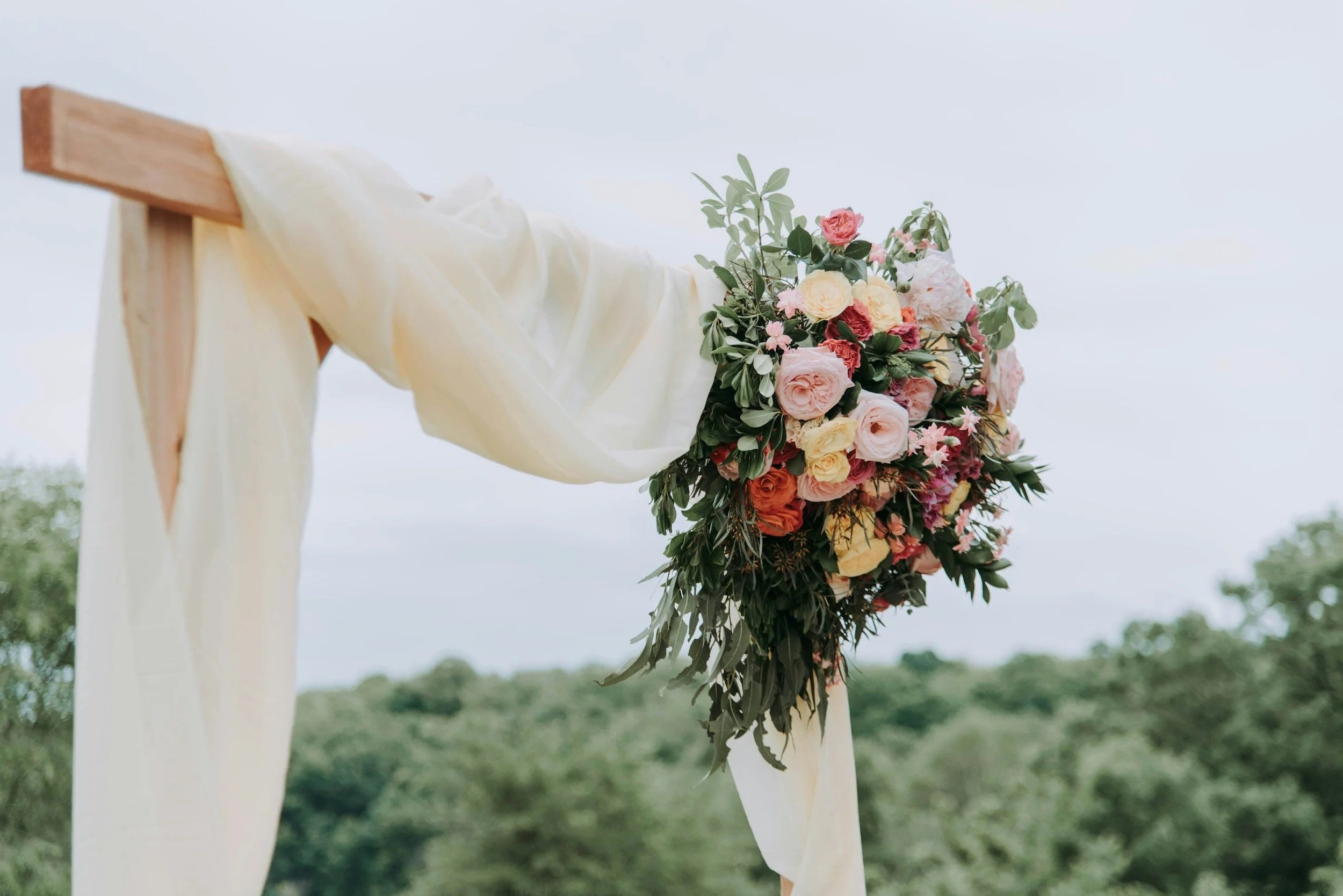 A decorated outdoor structure with cream fabric and a lush bouquet of pink, peach, and white flowers with green foliage against a cloudy sky and green landscape background.