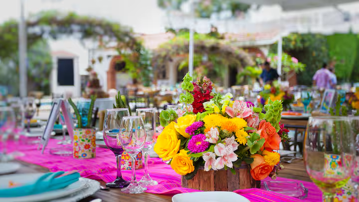 Colorful table setup with pink table runner, floral centerpiece with yellow, pink, and orange flowers, wine glasses, and place settings at an outdoor event or party