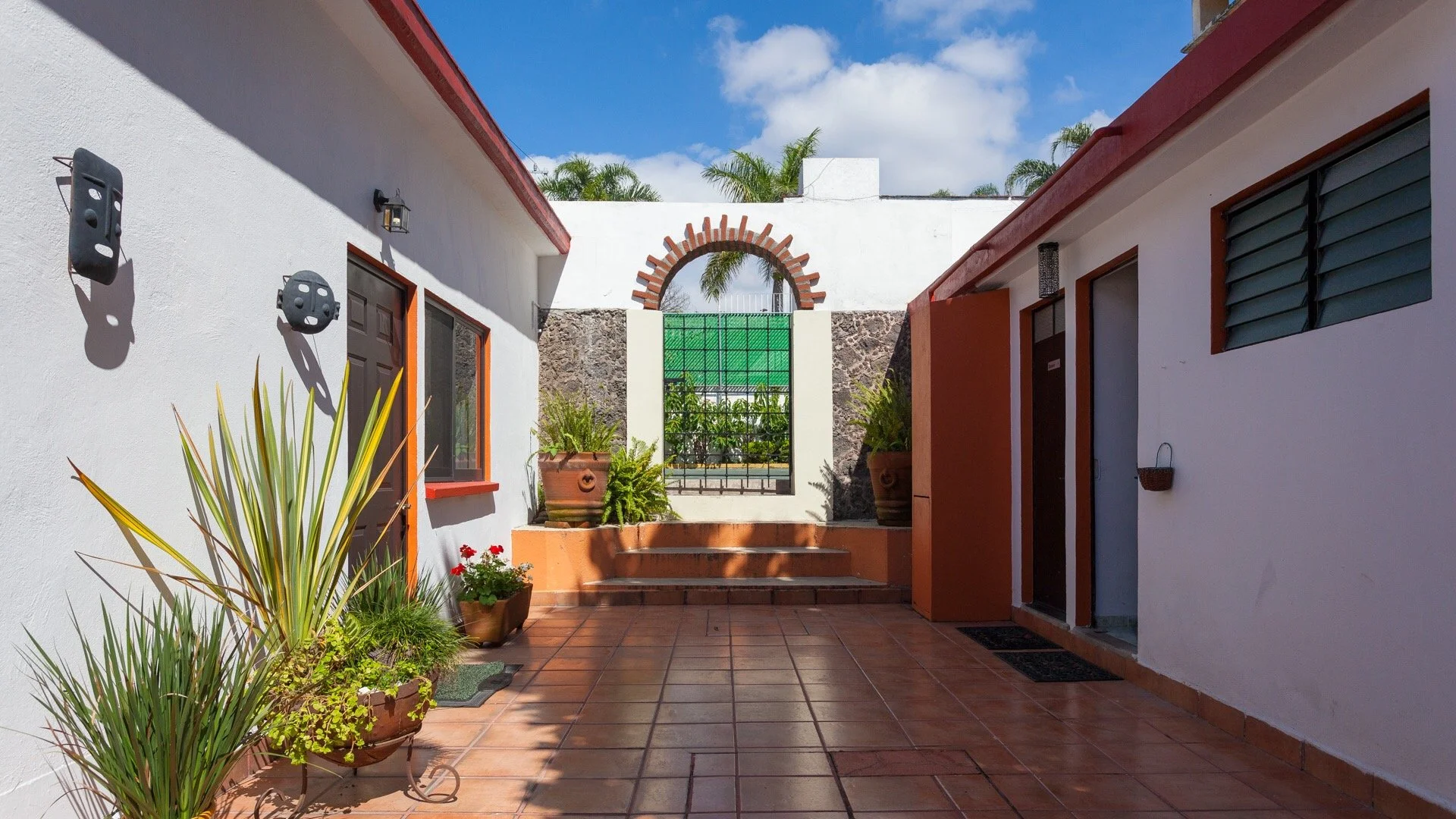 A courtyard with white walls, terracotta floor tiles, potted plants, and an arched brick decorative opening in the background. There are windows with shutters and a door on the walls.