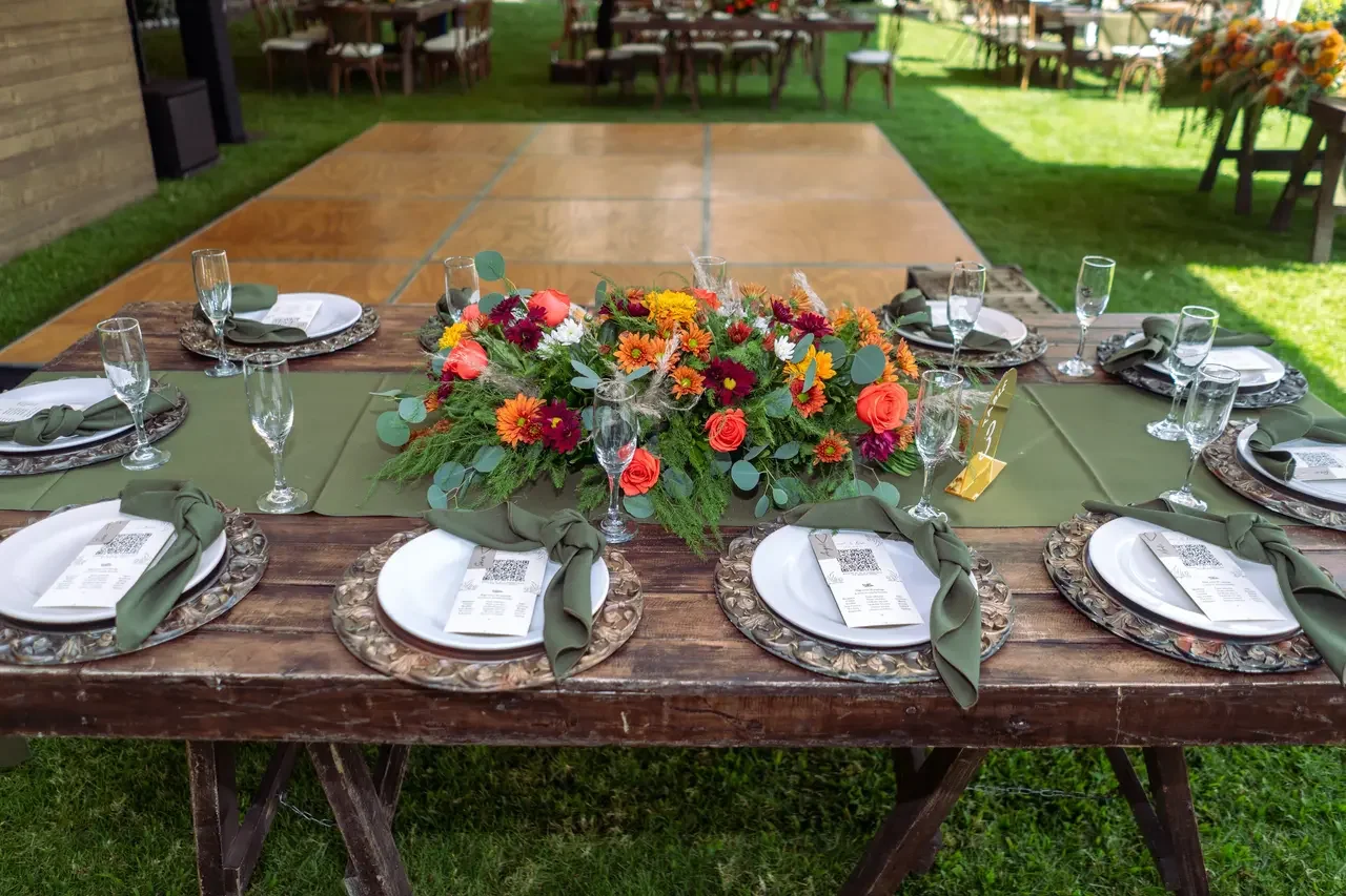 Outdoor banquet table set with floral centerpiece, green table runner, place settings with plates, napkins, and clear glasses, on a grass lawn with additional tables and chairs in the background.