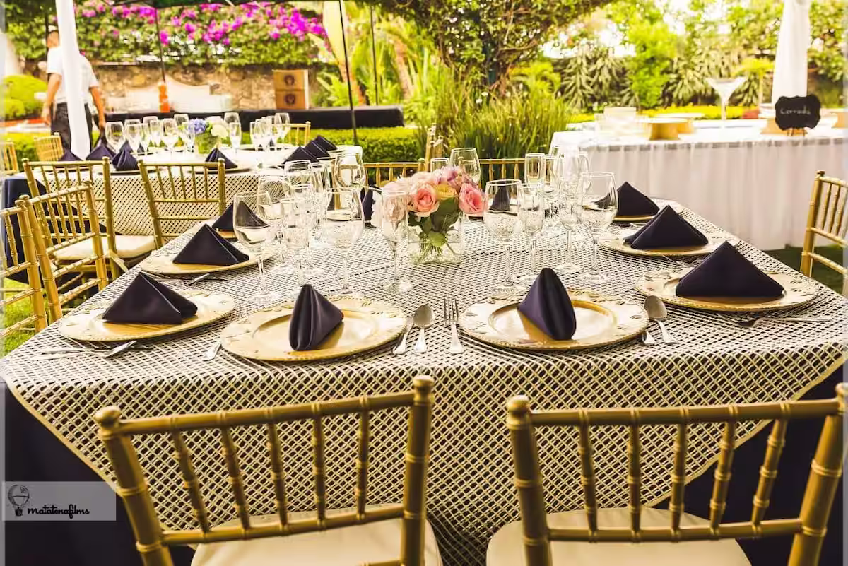 Elegant outdoor banquet table setup with gold Chiavari chairs, black napkins, wine glasses, and a floral centerpiece with pink flowers. Sunlit garden background.