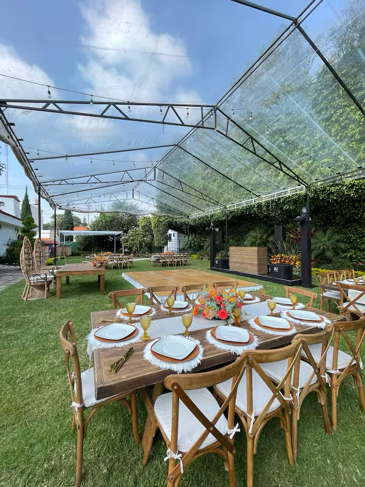 Outdoor event setup with a wooden table decorated with white placemats, golden glasses, and a floral centerpiece, under a clear tent on a grassy lawn with additional tables and a stage in the background.