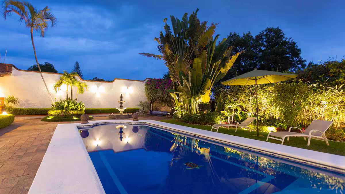Nighttime view of a backyard with a swimming pool, surrounded by greenery, palm trees, lounge chairs, umbrella, and garden lights