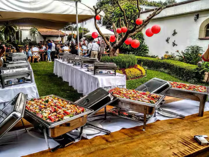 Buffet table with chafing dishes filled with food at an outdoor gathering, with guests in the background and decorative red lanterns hanging from a tree.
