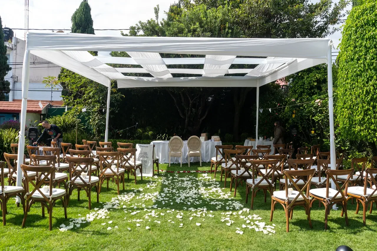 Outdoor wedding setup with chairs arranged on grass, flower petals forming an aisle, a canopy, and a table with chairs at the front, surrounded by trees and greenery.