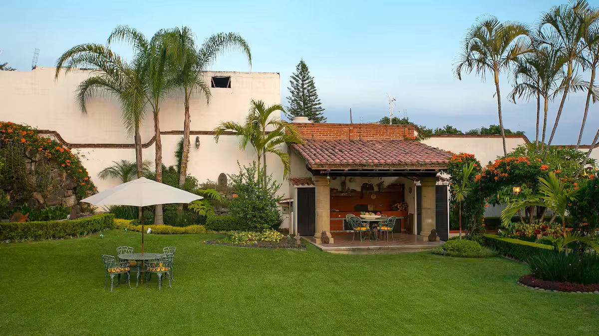 A backyard with a grassy lawn, outdoor dining table with chairs and a large umbrella, lush tropical plants, trees, and a small covered patio area with a table and chairs, set against a white wall and blue sky.