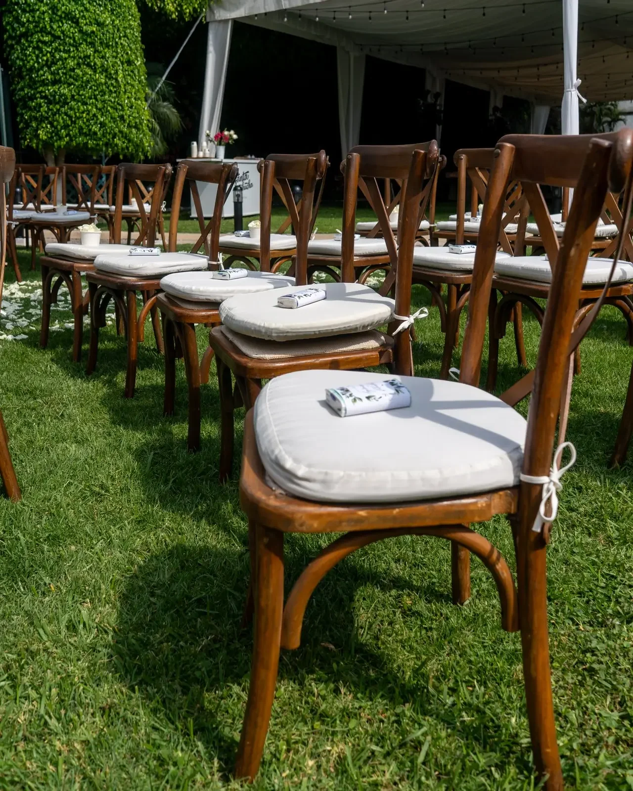 Empty outdoor wedding chairs with white cushions and napkins on green grass under a large tent.