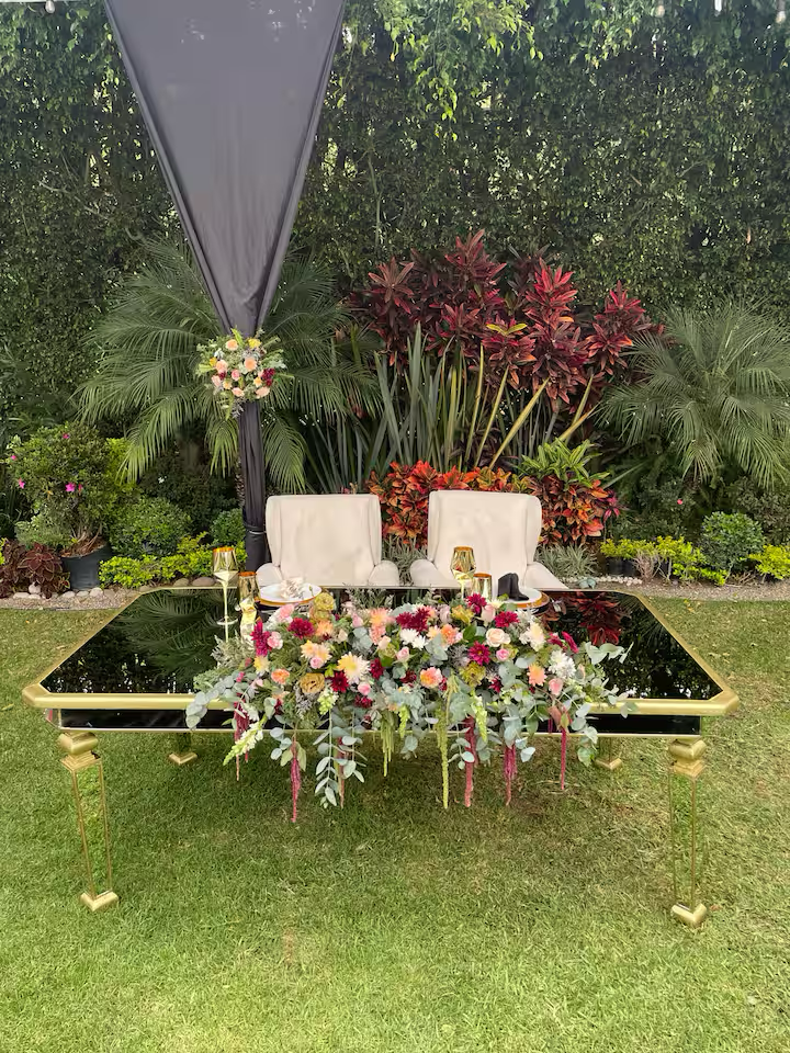 Elegant outdoor wedding or event setup with a mirrored table decorated with a colorful flower arrangement, two white chairs, and gold tableware, set in a lush garden with green foliage and vibrant plants.