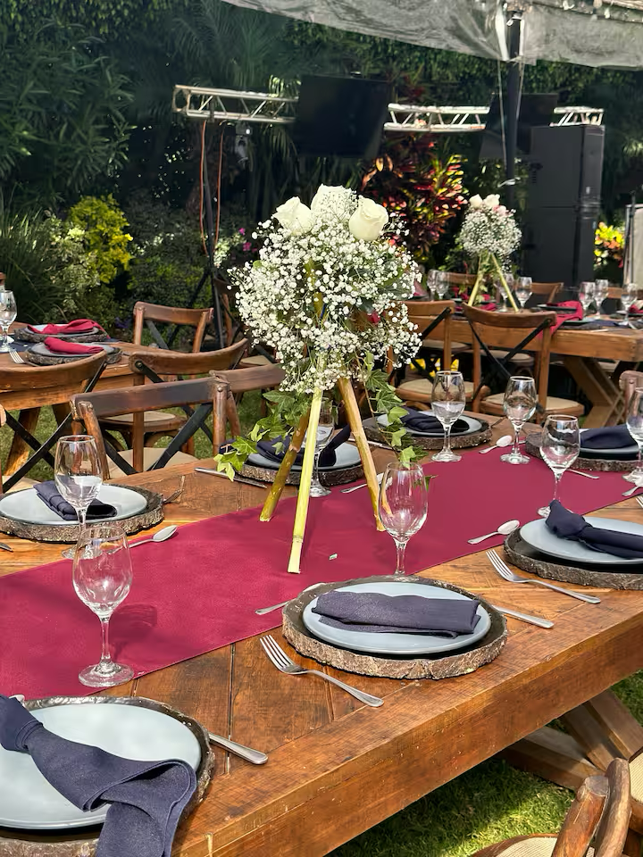 Outdoor banquet table decorated with a burgundy table runner, black napkins, white plates with wooden slice chargers, and clear wine glasses. A floral centerpiece with white roses, baby’s breath, and greenery is placed on a tripod stand.