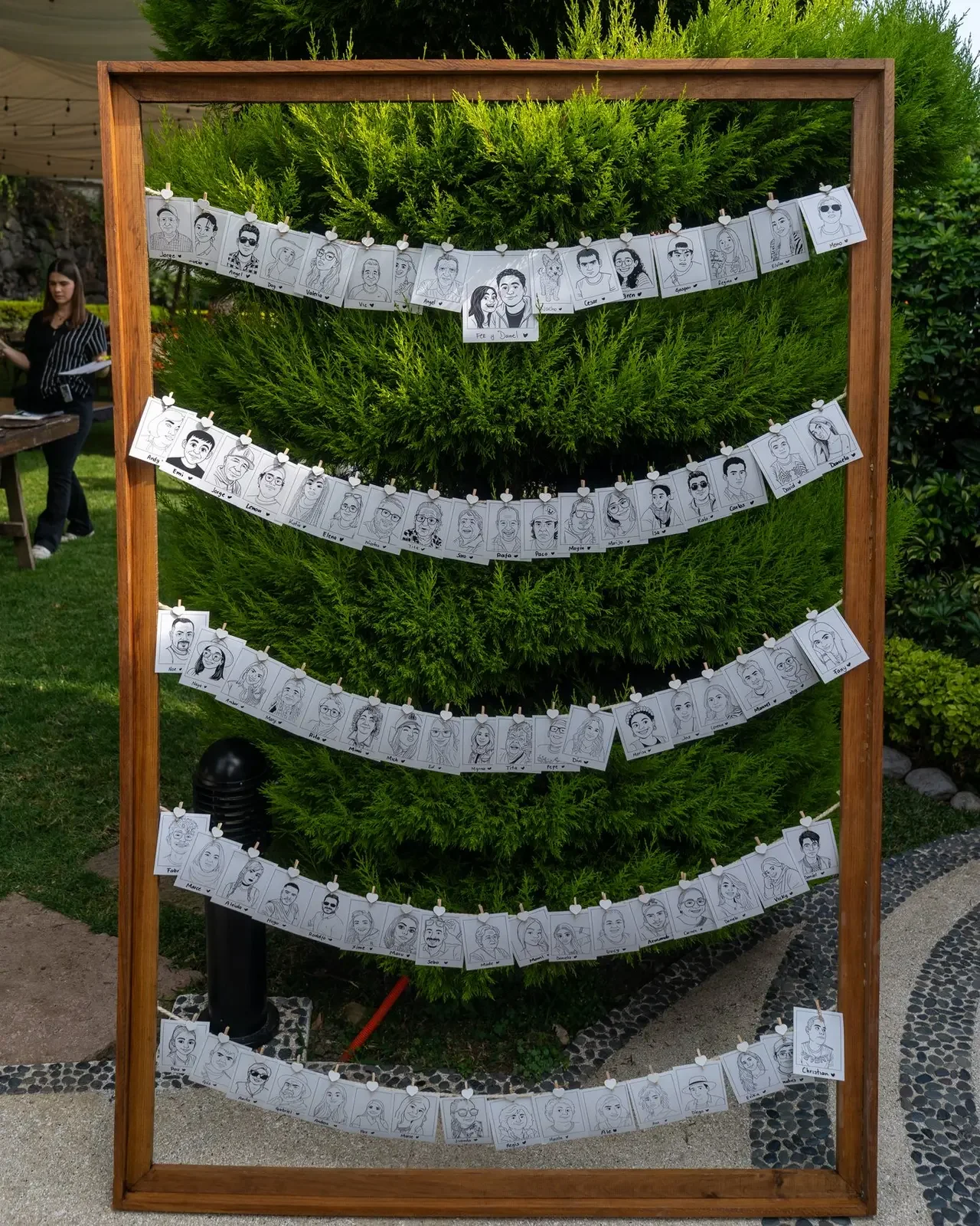 Display board with hand-drawn caricatures of people, hung on strands with clothespins against a green bush background, at an outdoor event.