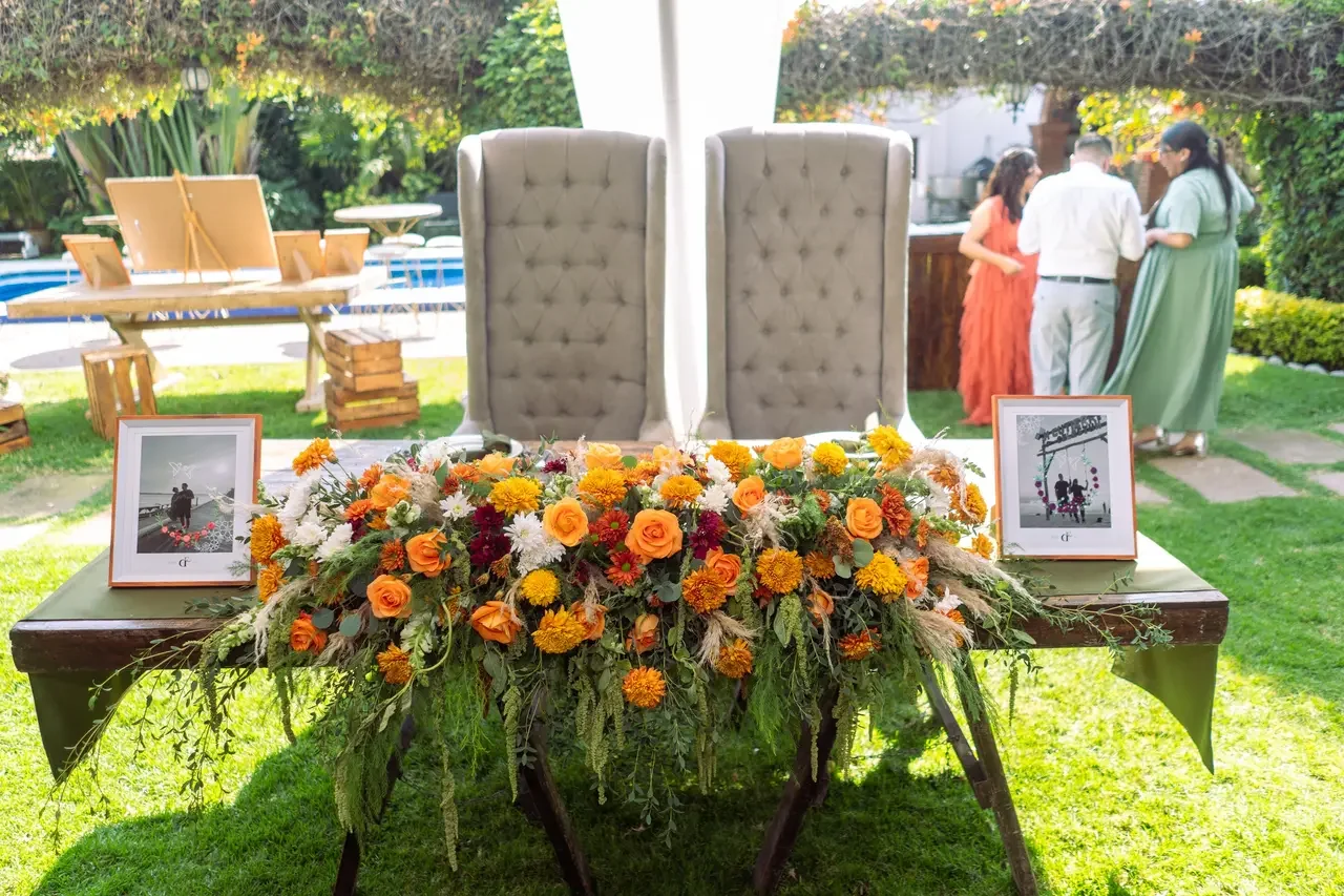 Decorative table with a large flower arrangement of orange, yellow, and white flowers, framed photographs on each side, and two high-backed beige chairs behind the table, set outdoors during daytime with people conversing in the background.