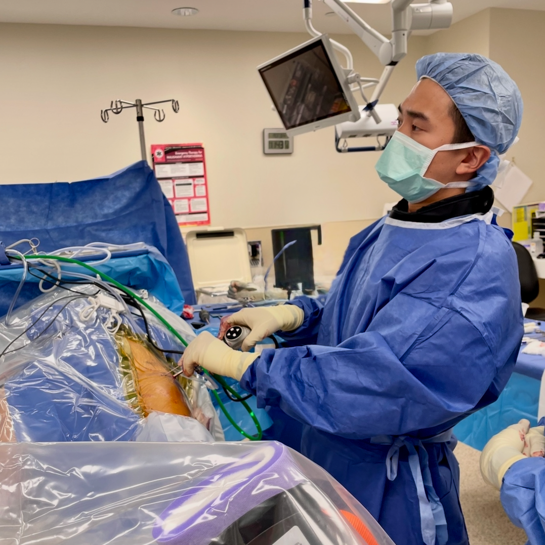 Surgeon in blue scrubs, mask, and cap performing surgery with medical instruments and monitoring equipment in an operating room.