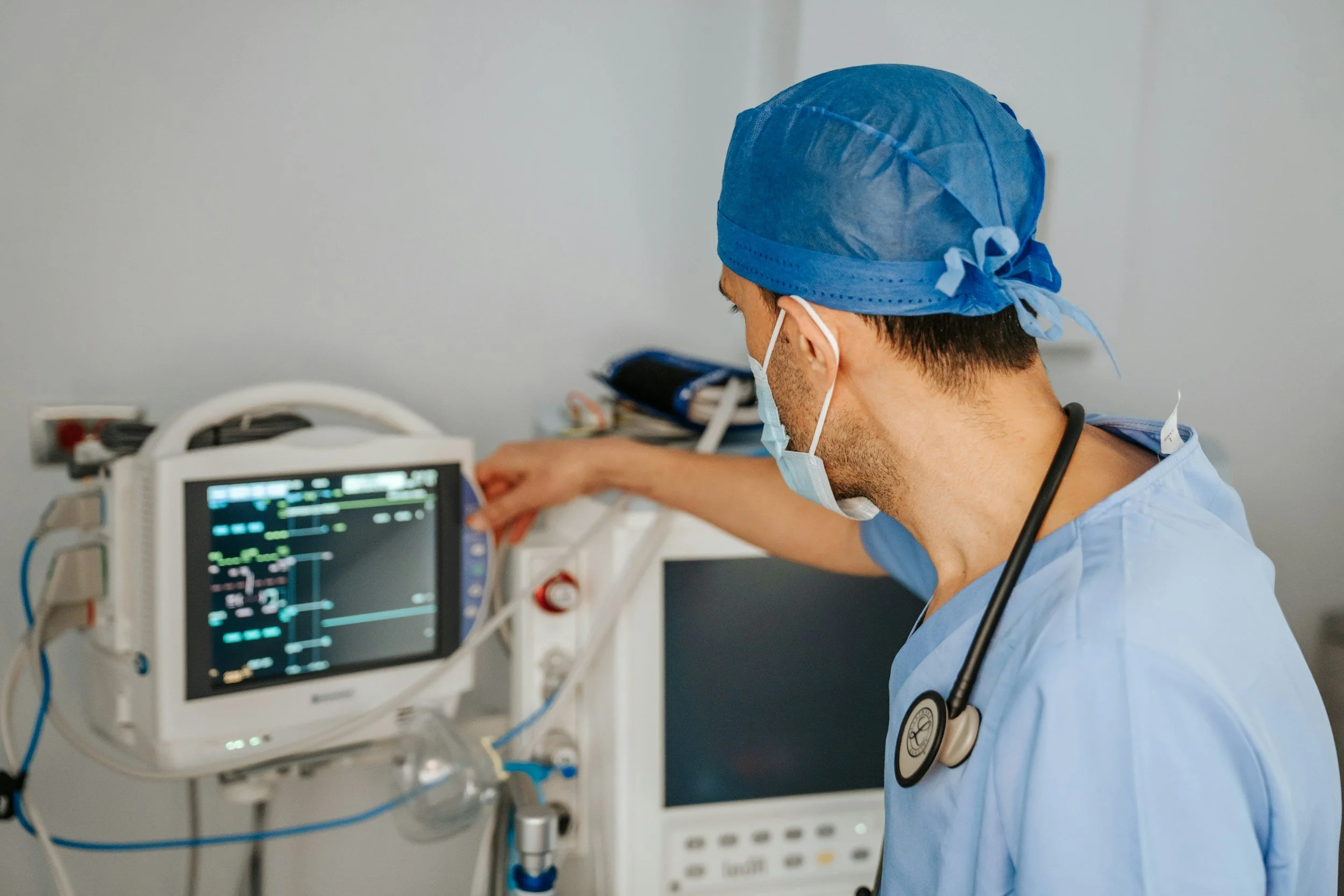A healthcare professional wearing blue scrubs, a blue surgical cap, and a mask is working with medical monitoring equipment in a hospital or clinic setting. The professional appears to be adjusting or checking a medical device with a screen showing vital signs.