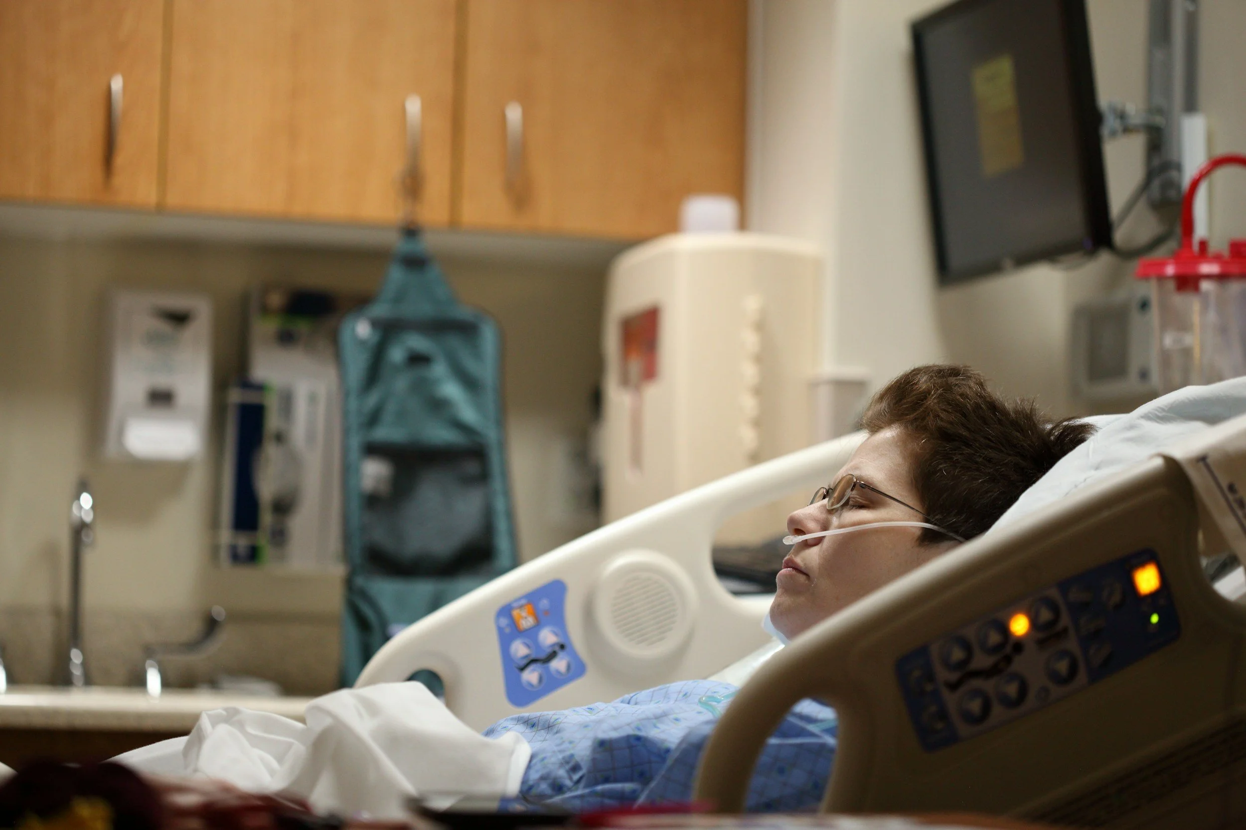 Woman lying in hospital bed with head turned to the side, wearing glasses and a nasal oxygen tube. Medical equipment and wooden cabinets are visible in the background.