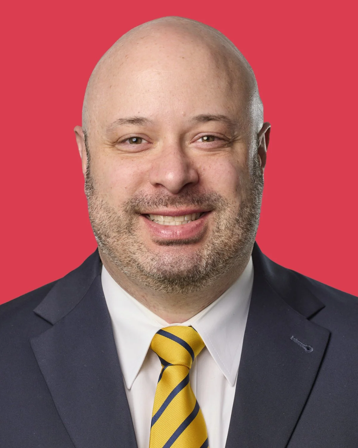 A man with a bald head and a beard, wearing a dark suit, white shirt, and yellow striped tie, smiling against a solid red background.