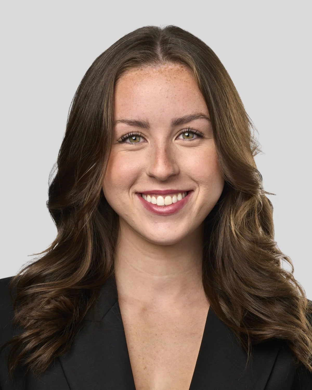 A young woman with long, wavy brown hair smiling and wearing a black blazer against a light gray background.