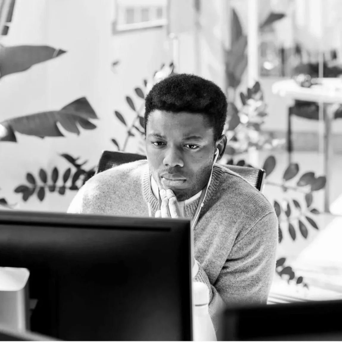 A young man with short hair looking at a computer screen, wearing headphones, in an office with plants in the background.
