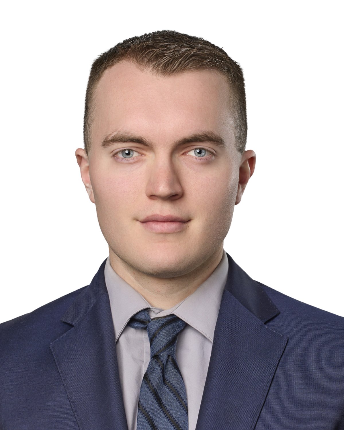 A young man with short brown hair, blue eyes, wearing a navy blue suit, light gray shirt, and striped tie, posing against a white background.