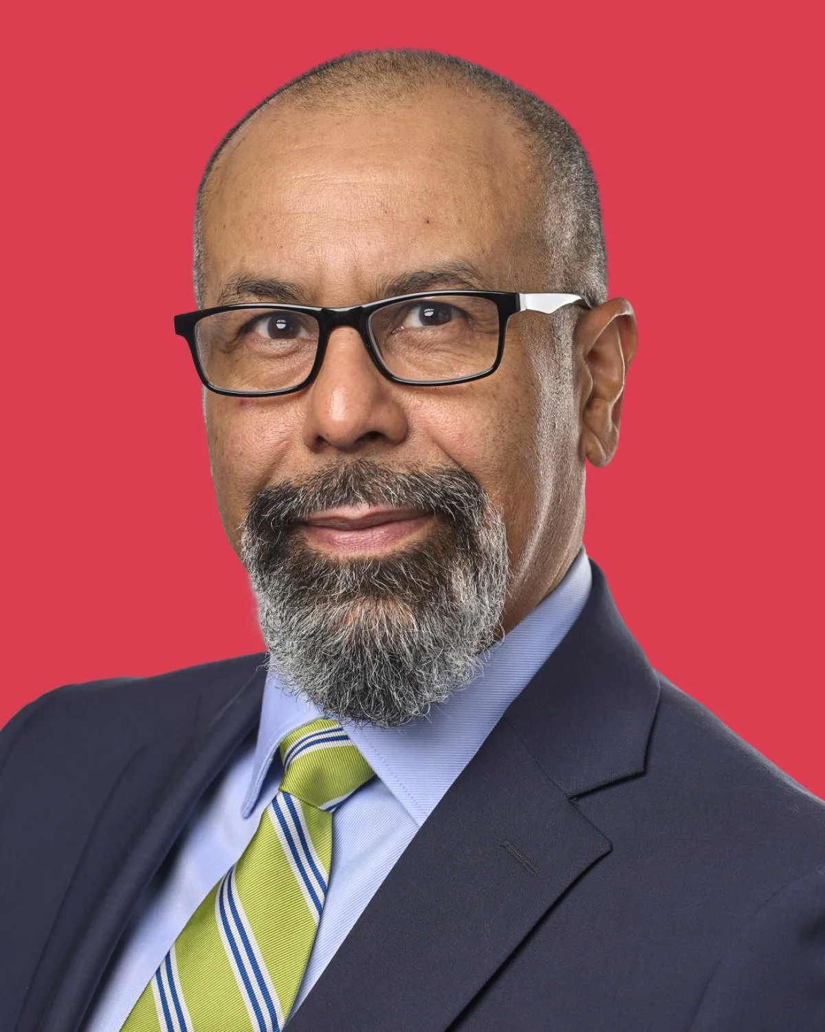 A middle-aged man wearing glasses, a dark suit, light blue shirt, and yellow-green striped tie, with a bald head and gray goatee, posing against a solid red background.