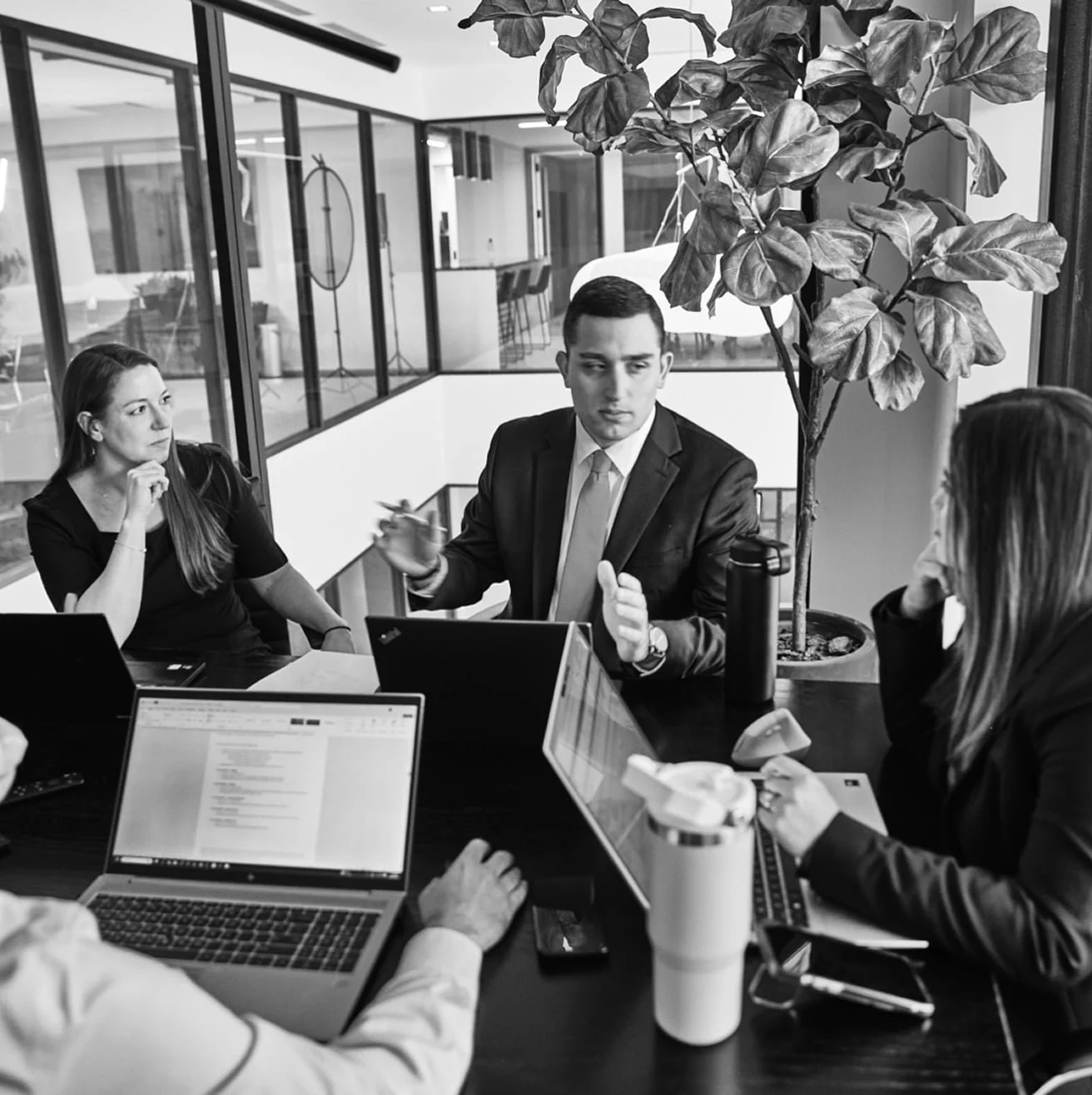 A group of colleagues in business attire having a meeting around a conference table with open laptops.
