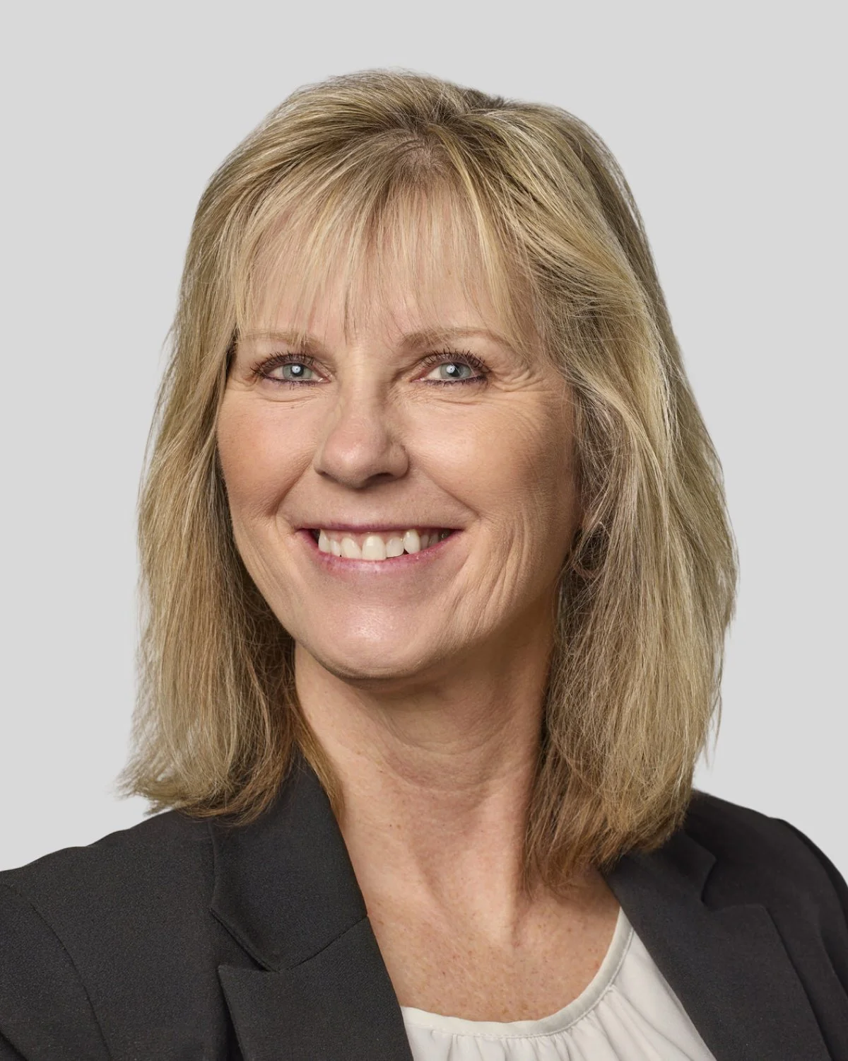 Portrait of a middle-aged woman with blonde hair, blue eyes, wearing a black blazer and smiling, against a plain light gray background.