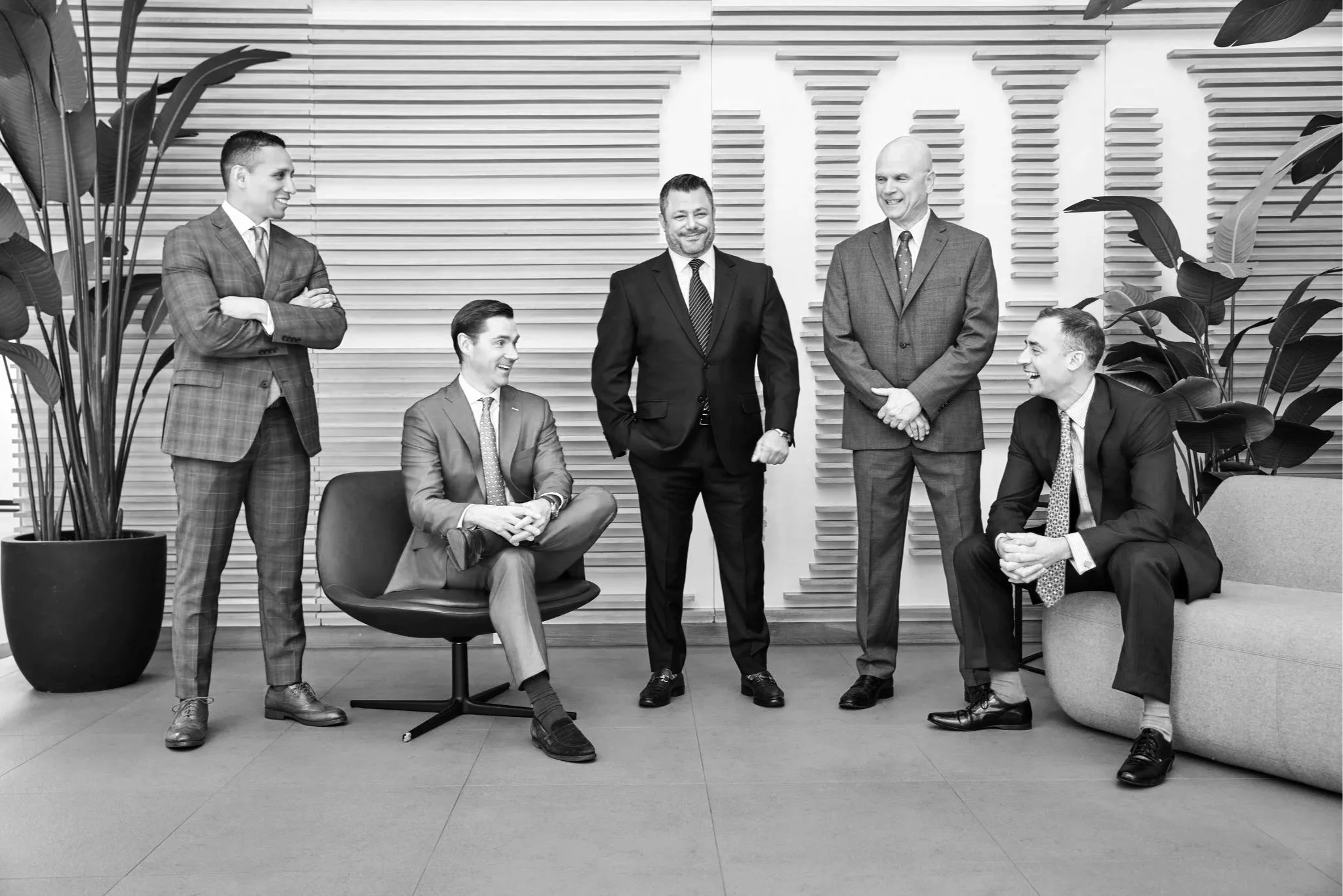 Black-and-white photograph of six businessmen in a modern office setting. They are engaged in conversation and appear to be enjoying themselves. Three men are seated, while three are standing. The background features a textured wall with horizontal wooden slats, and there are large potted plants on each side of the group.