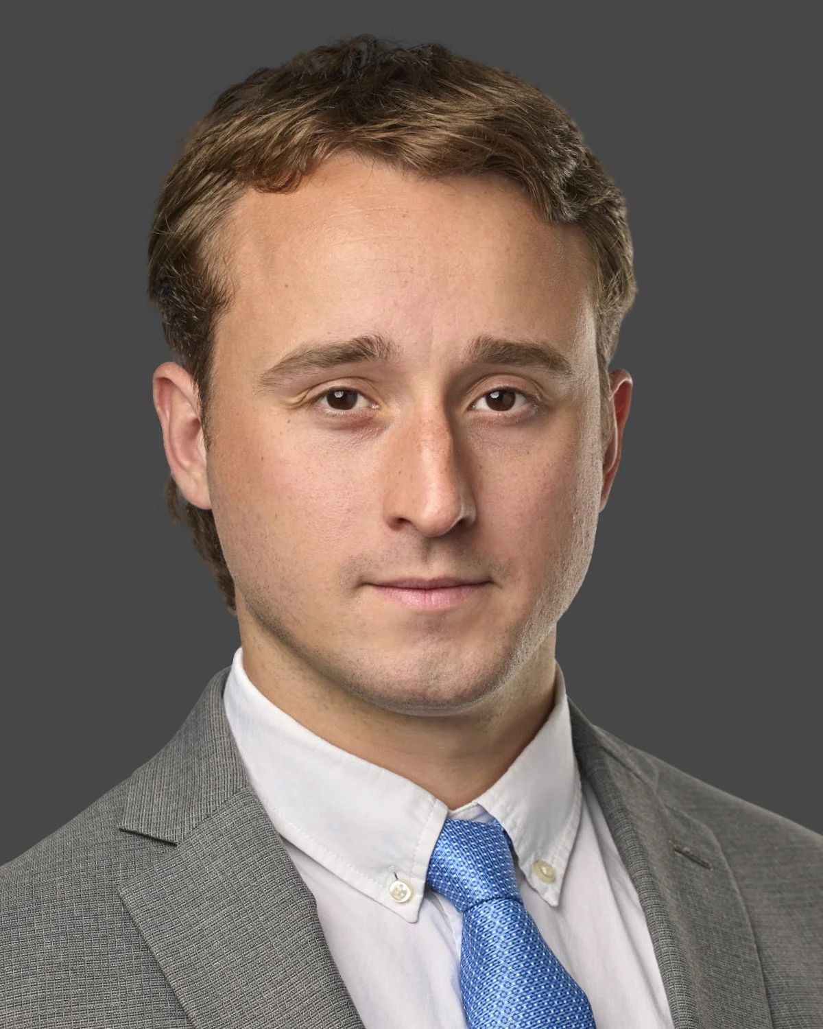 Portrait of a young man with light brown hair, wearing a gray suit, white shirt, and blue tie, against a plain gray background.