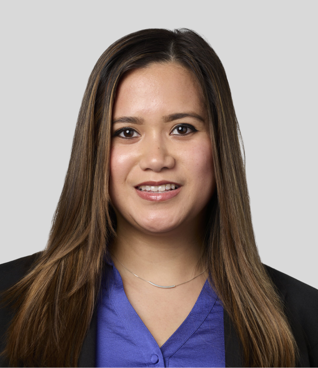 Professional headshot of a young woman with long brown hair, wearing a blue blouse and black blazer, smiling against a light gray background.