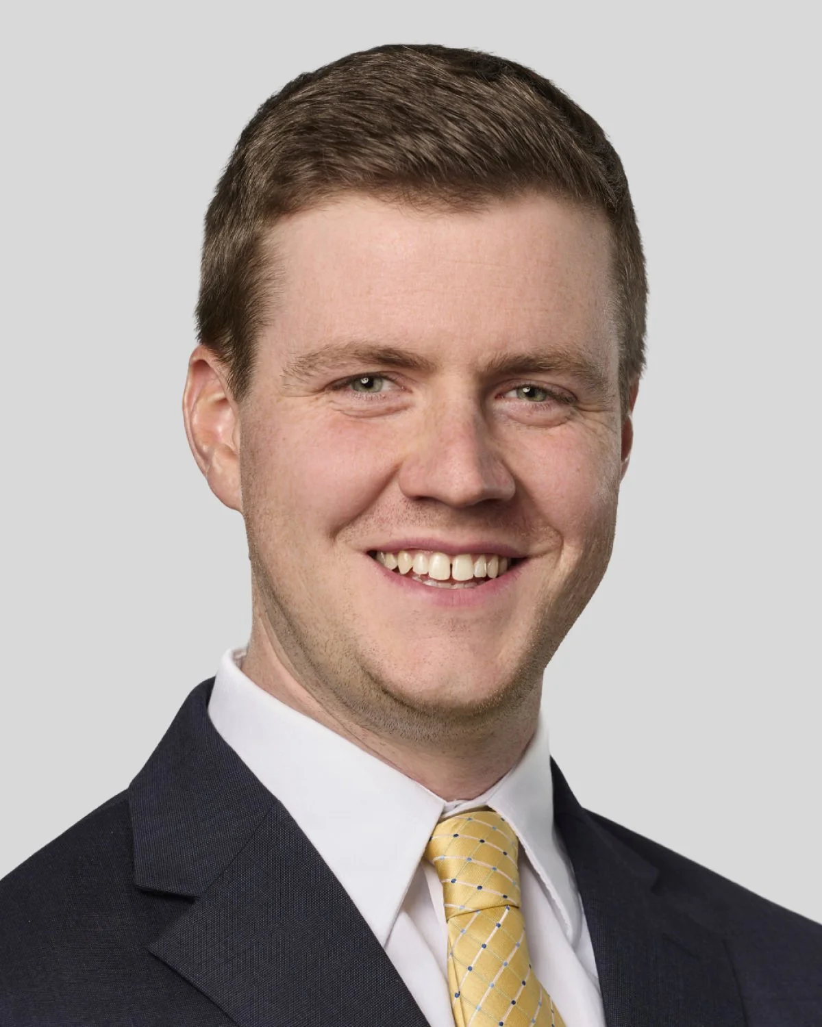 Headshot of a smiling man with short brown hair, wearing a black suit, white shirt, and yellow tie, against a light gray background.