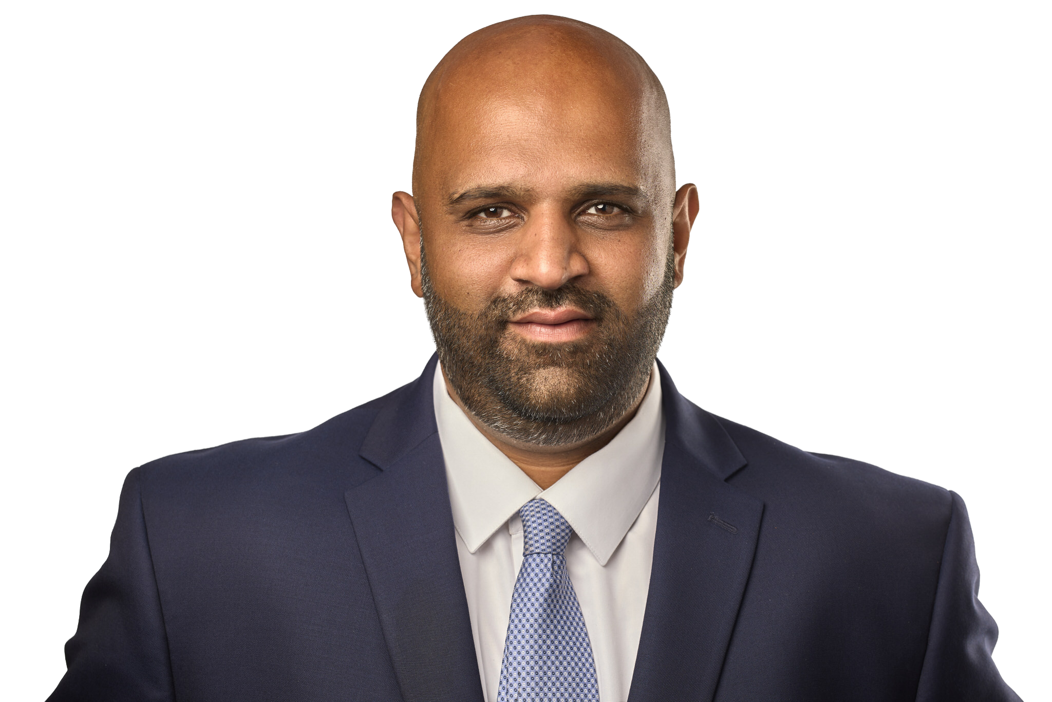 A man in a dark suit, white shirt, and light blue tie, smiling confidently, with a clean-shaven head and beard, against a plain dark background.