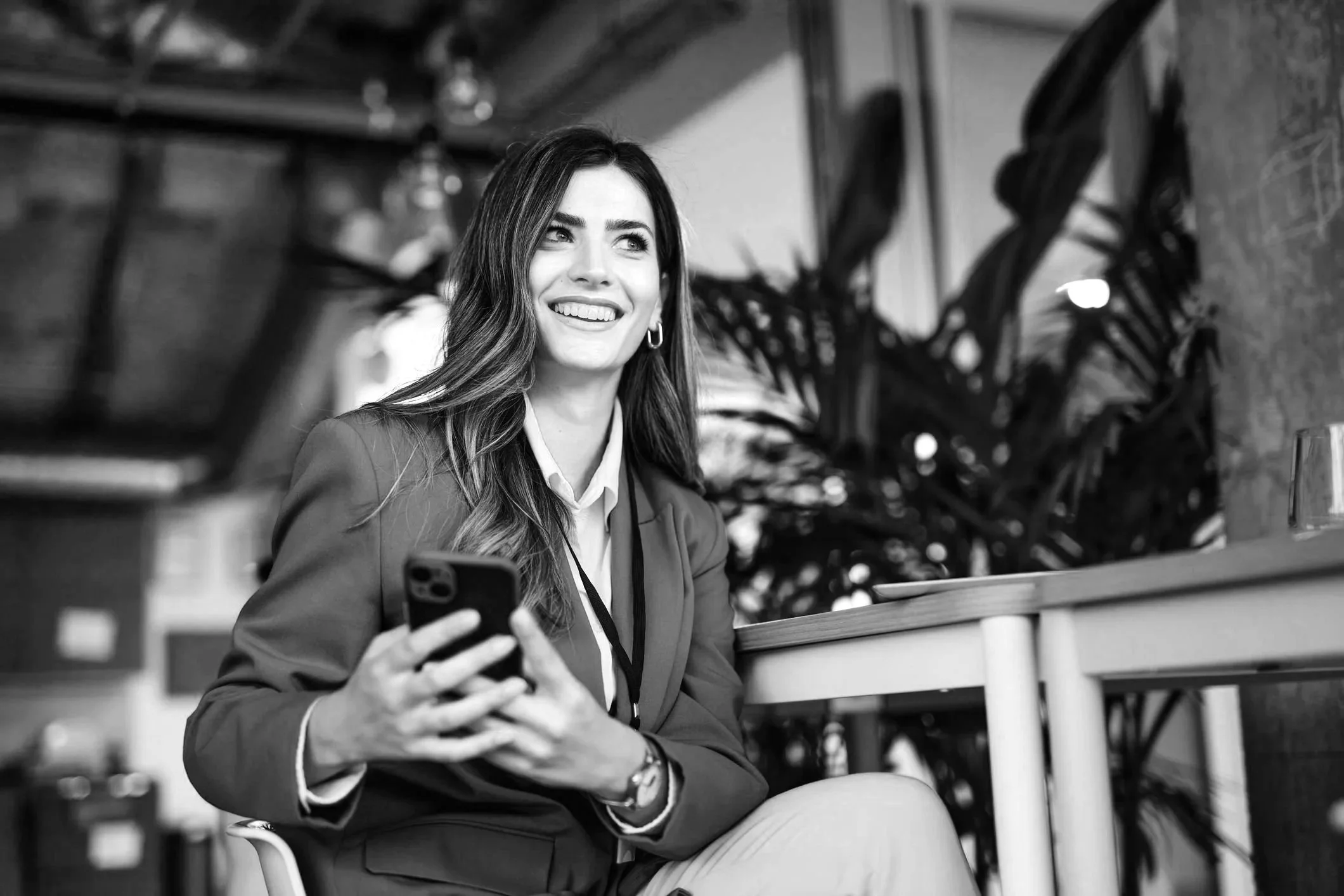 A smiling woman with long hair, wearing a blazer over a blouse, sitting at a table with a smartphone in her hand, in a modern indoor setting.