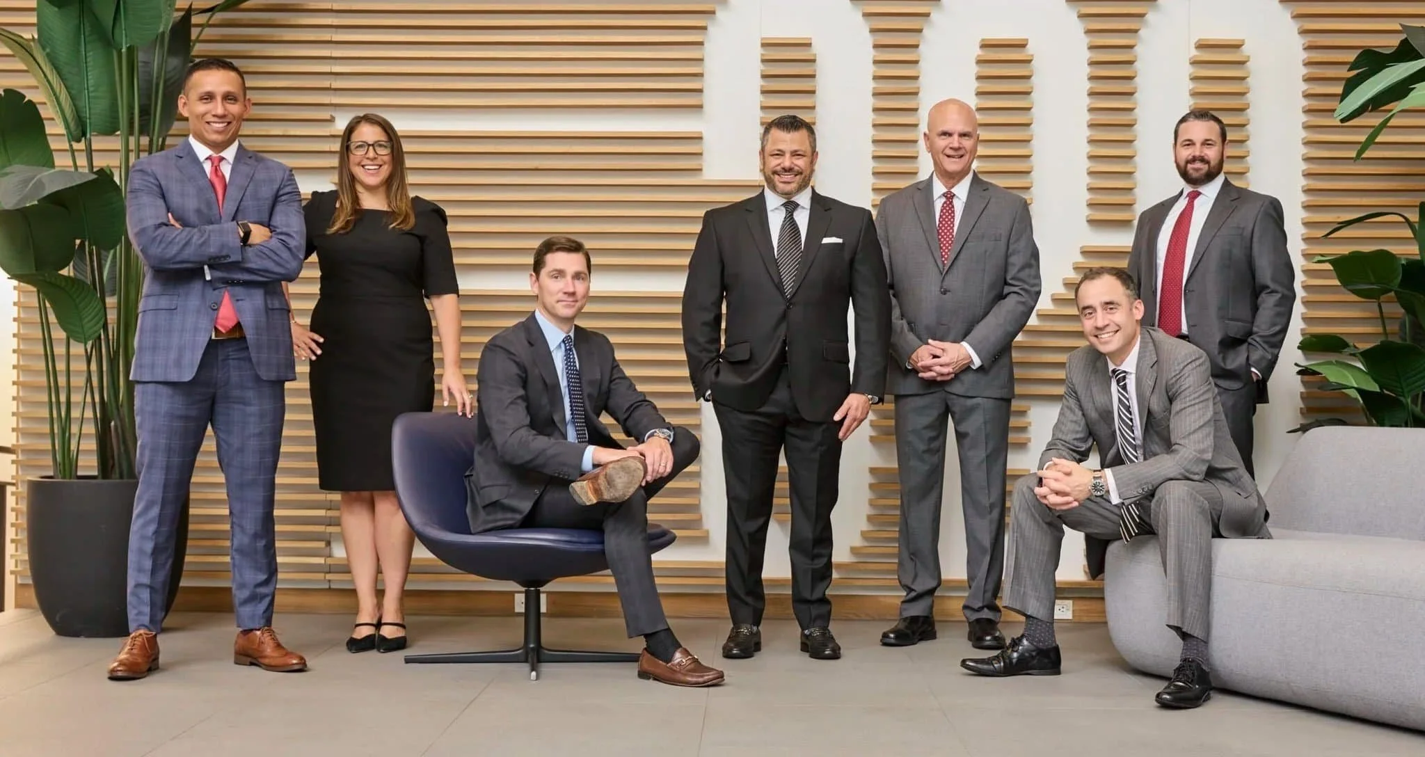 A group of nine professionals, seven men and two women, posing together in a modern office with decorative wooden wall panels and large potted plants. Some are standing and others are sitting, all dressed in business attire.