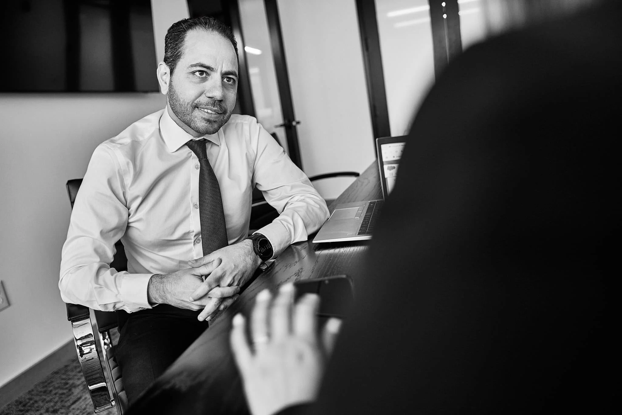 A man with dark hair, beard, wearing a white shirt, dark tie, and a smartwatch, sitting at a conference table in a meeting room, engaged in conversation.