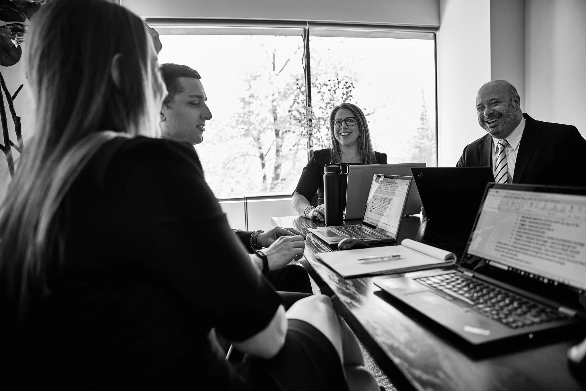 A group of professionals in a business meeting in a modern office, with one woman smiling and a man in a suit, working on laptops and documents.