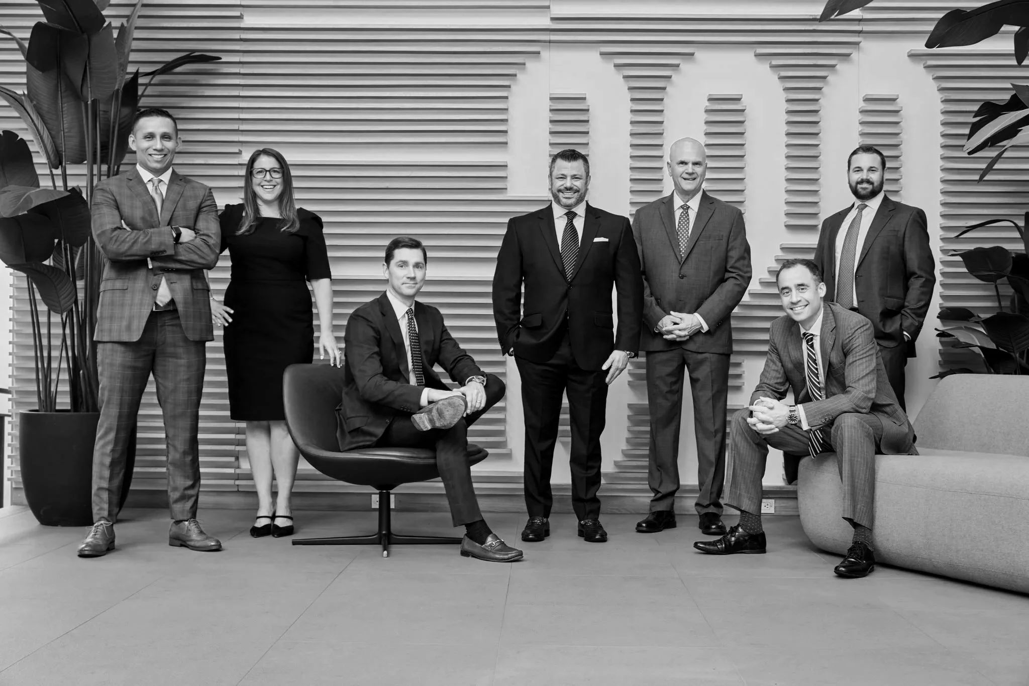 Group of nine diverse professionals in business attire in an office lobby with wooden wall decor and large plants, smiling for a group photo.