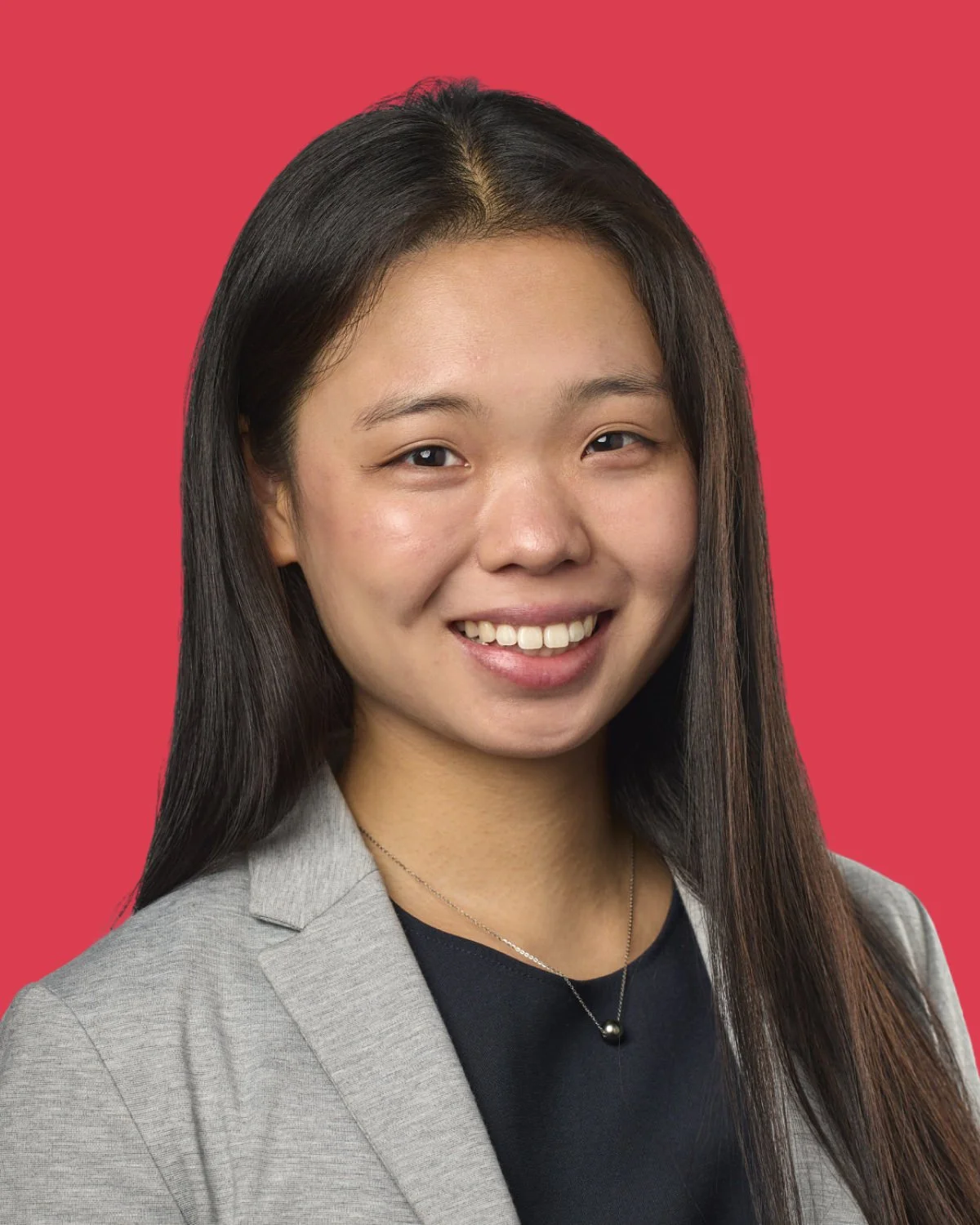 Headshot of a young Asian woman with long dark hair, smiling, wearing a gray blazer over a black top, against a coral background.
