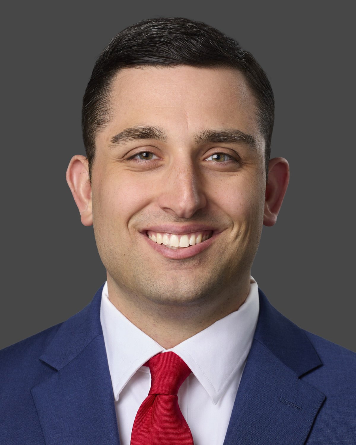 Professional headshot of a smiling young man with dark hair, wearing a blue suit, white shirt, and red tie, against a gray background.