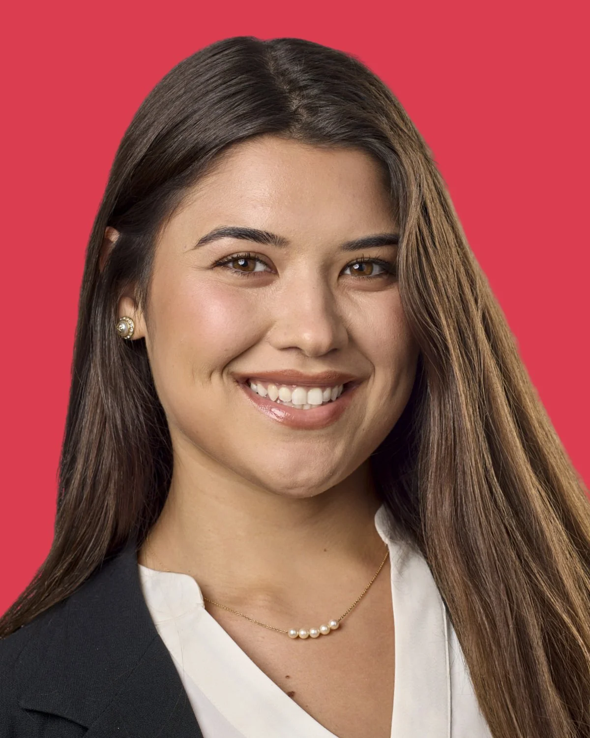 A young woman with long brown hair, smiling, wearing pearl earrings and a pearl necklace, dressed in a black blazer and white blouse, against a pink background.