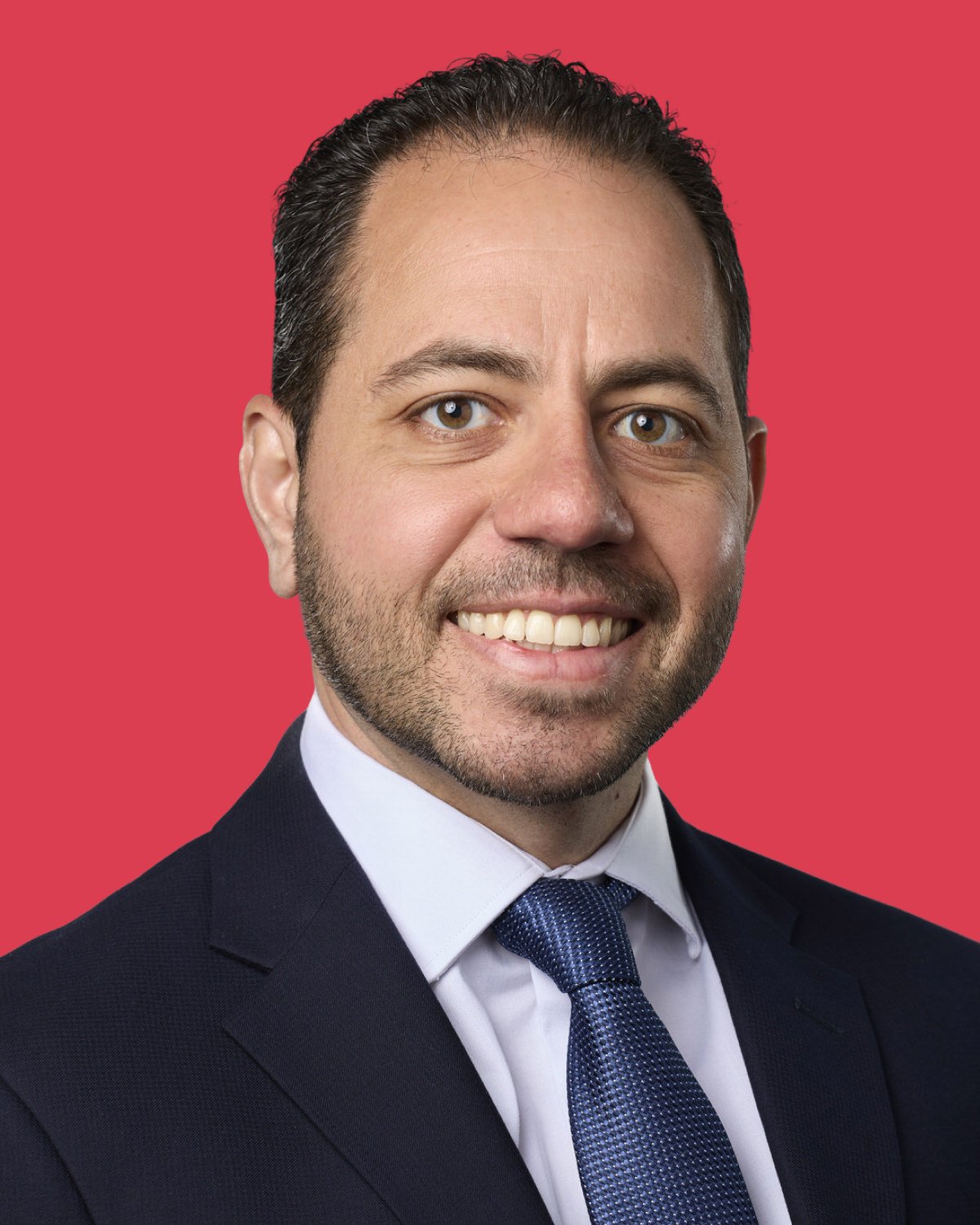 A professional headshot of a man wearing a dark suit, white shirt, and blue tie, smiling against a red background.