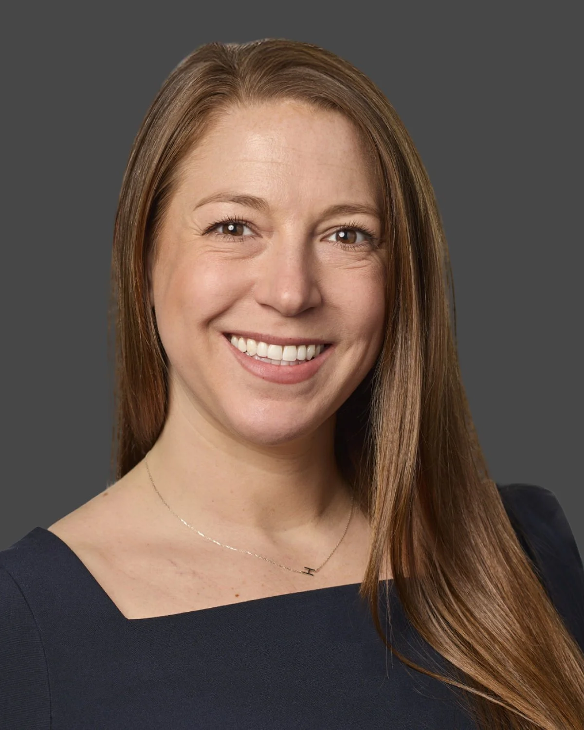 A headshot of a smiling woman with long, straight, auburn hair wearing a dark dress and a delicate necklace, against a gray background.